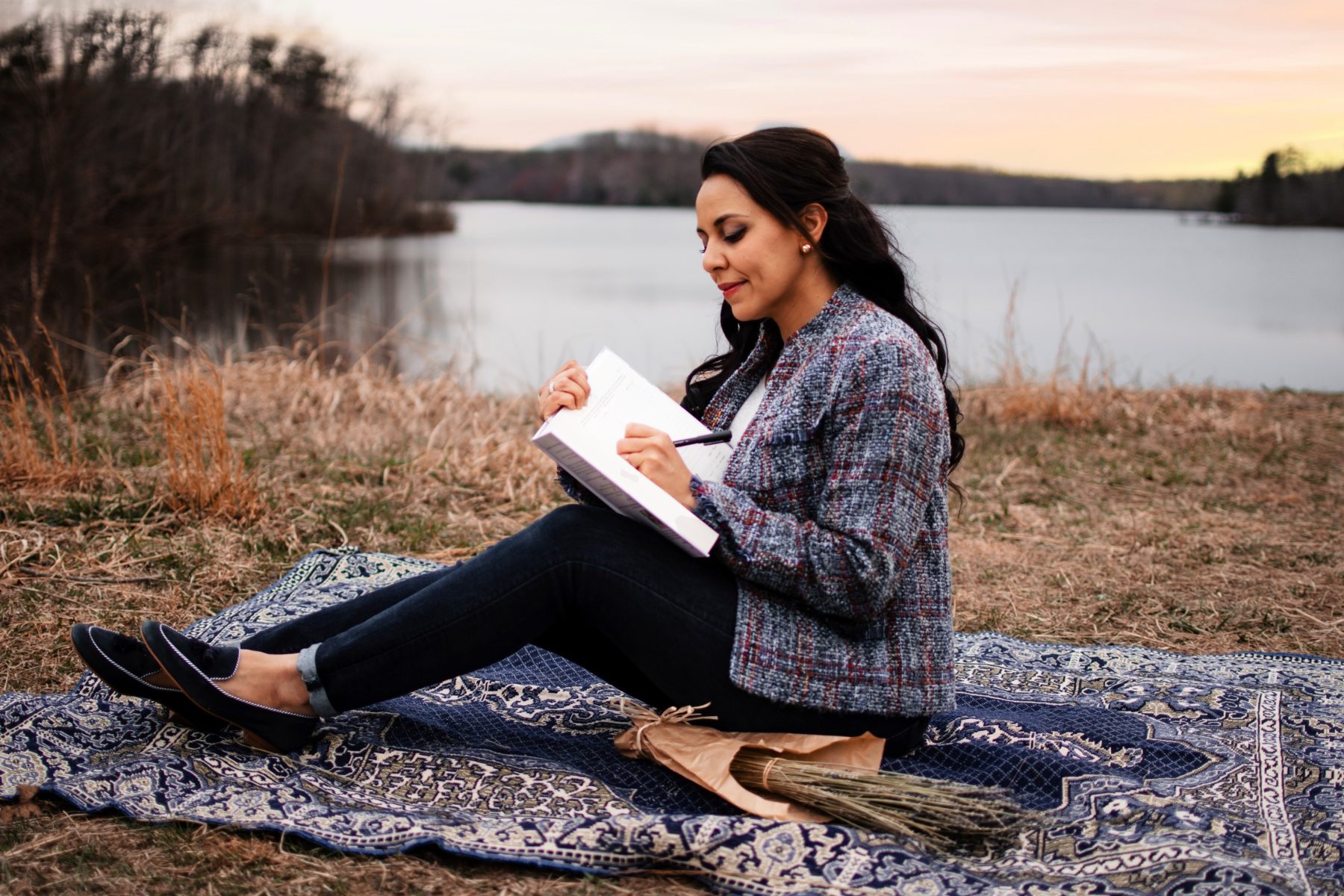 A woman journaling at the edge of a lake at sunset, seated on a Persian rug in golden dry grass